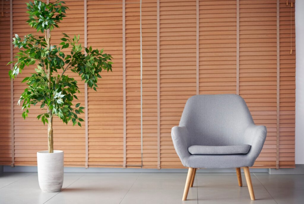 A modern gray armchair with wooden legs sits next to a potted artificial plant against a backdrop of wooden blinds, enhancing a contemporary interior.